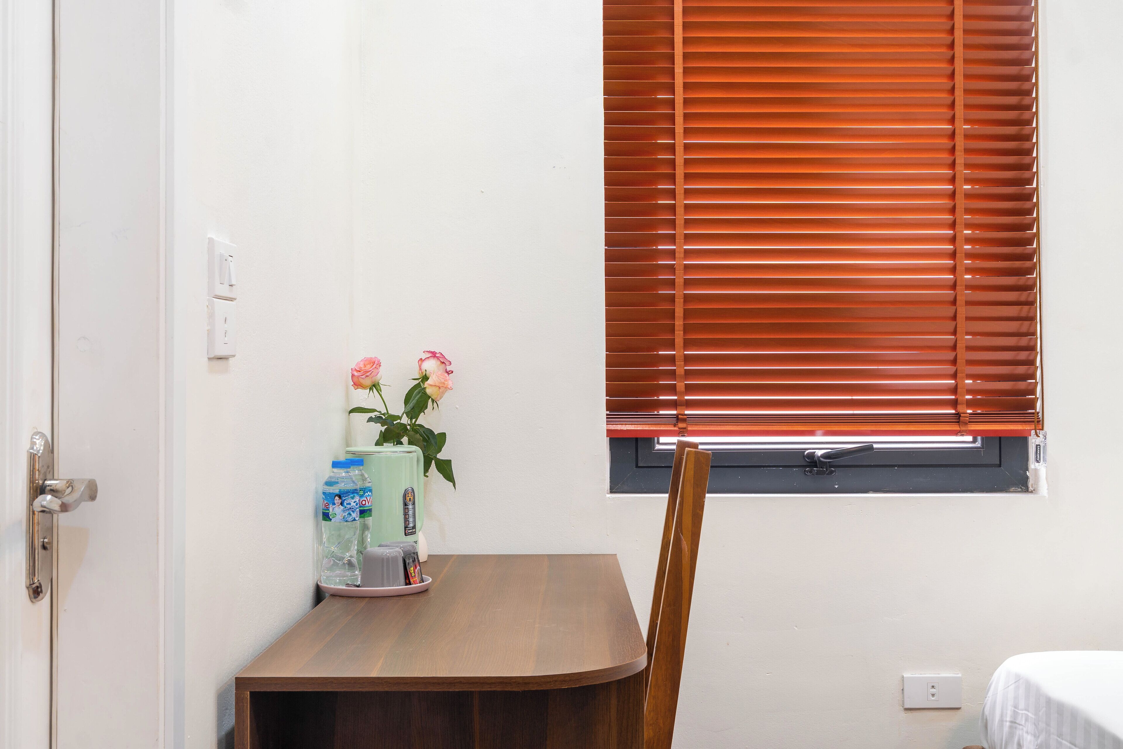 A room with a wooden table, a chair, a window with blinds, and a wall-mounted light switch.