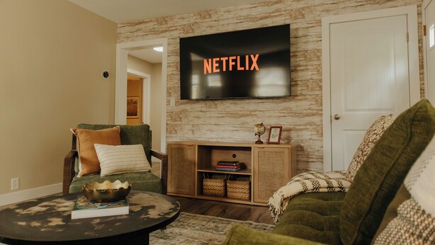 Un salon avec un téléviseur à écran plat fixé au mur, affichant le logo « NETFLIX », un meuble en bois en dessous et un canapé vert.