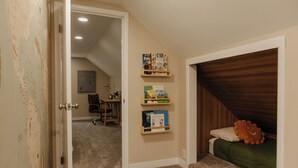 Une chambre avec un plafond en pente, une couvre-lit verte et une tête de lit en bois. On y trouve des étagères remplies de livres pour enfants et un petit bureau avec une chaise.