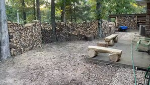 Een rustieke buitenruimte met banken en een stenen muur, omringd door bomen en een stapel hout.