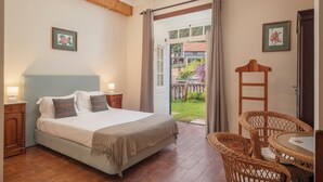 Ein Schlafzimmer mit einem Bett, Nachttischen, einem Korbstuhl, einem kleinen Tisch und Blick ins Freie.