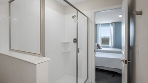 Une chambre d’hôtel moderne dotée d’une douche en verre, d’un lit donnant sur la ville et d’une porte menant à une autre chambre.
