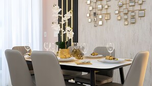 A modern dining area with a white table set for four, surrounded by grey chairs. A decorative wall with geometric patterns and a vertical gold strip is visible behind the table.
