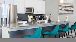 A modern kitchen with white cabinets, stainless steel appliances, and teal bar stools.