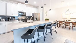 Uma cozinha moderna com armários brancos, uma ilha grande com banquetas, eletrodomésticos de aço inoxidável e iluminação pendente.