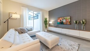 Uma sala de estar moderna com um sofá branco, uma mesa de centro, um abajur, uma TV fixada na parede e uma grande janela com cortinas.