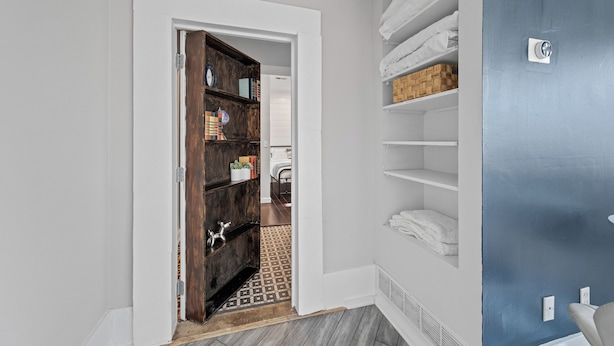 Un couloir avec une porte en bois donnant sur une chambre. Des étagères contenant des serviettes et une petite plante se trouvent sur la droite.