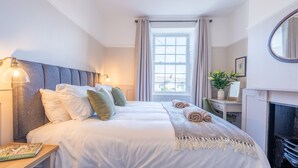 A neatly made bed with white linens and multiple pillows, a bedside lamp, a small table with a book, a window with curtains, and a fireplace with a mirror above it. - Church House - sleeps 6 guests  in 3 bedrooms (Beaumaris)