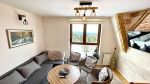 A modern living room with a sectional sofa, a round coffee table, and a large window offering a view of trees and mountains. - NPL Gorstak apartments (Jahorina)