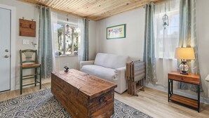 Un salon chaleureux avec un plafond en bois, un fauteuil vert, un canapé blanc, une commode en bois et une lampe sur une table d’appoint.