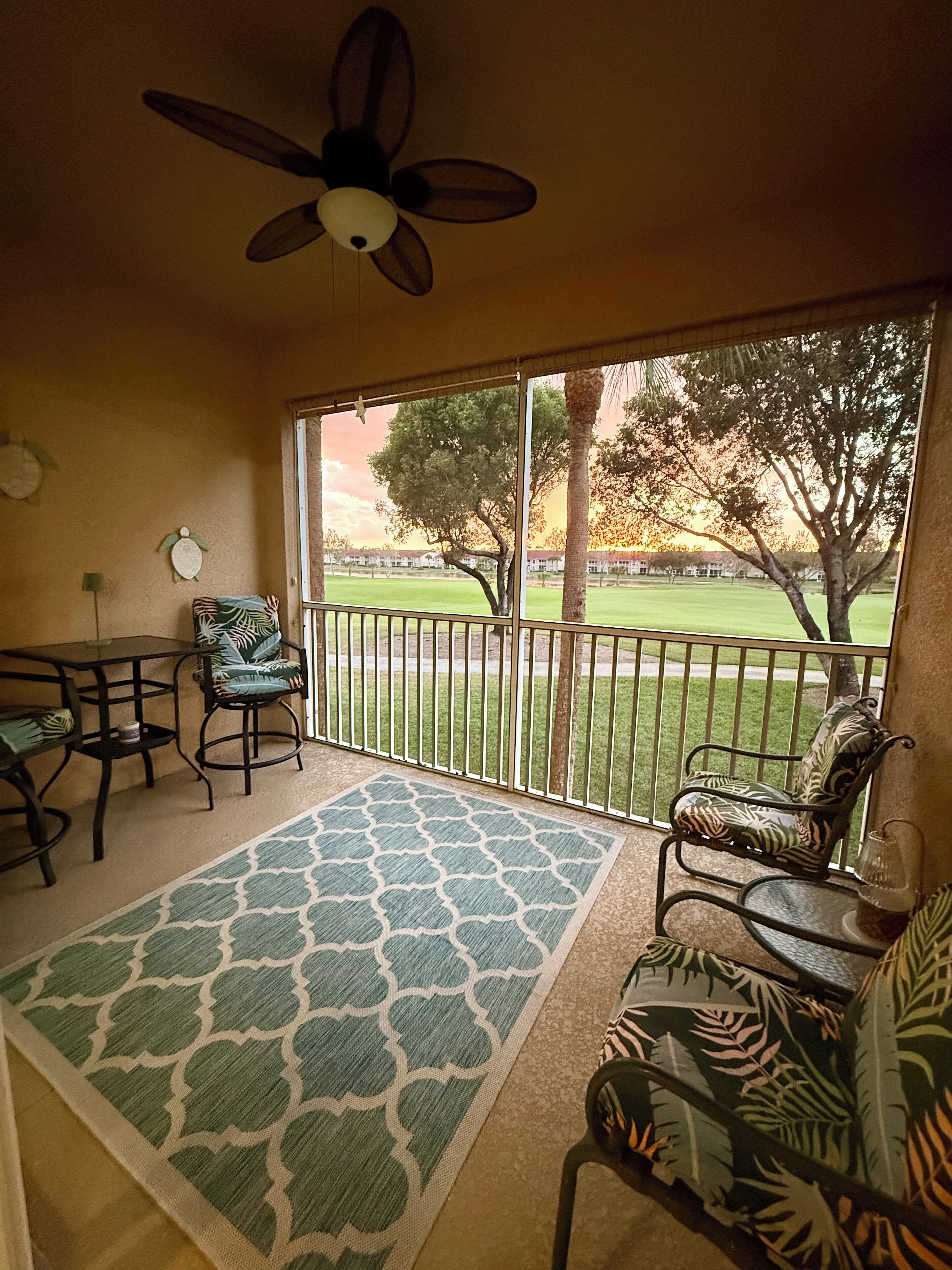 A room with a ceiling fan, a view of a green lawn, and a patio area with chairs and a table.