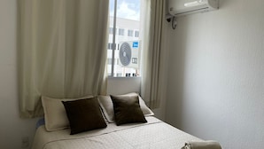 Une chambre d’hôtel avec un lit, des rideaux blancs, une fenêtre donnant sur des immeubles et un climatiseur fixé au mur.