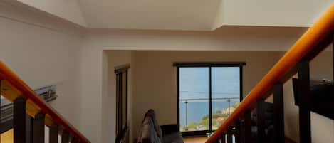 A spacious room with a large window offering a view of the sea, a wooden staircase, and a modern chair.