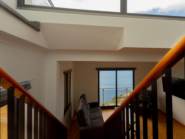 A spacious room with a large window offering a view of the sea, a wooden staircase, and a modern chair.