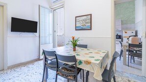 Uma área de refeições com mesa, cadeiras, uma planta em vaso, uma TV de tela plana e um quadro na parede.