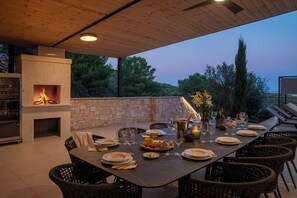 Un espacio de comedor al aire libre con pared de piedra, chimenea integrada, sillas de mimbre y una mesa dispuesta para una comida.