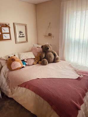 A bedroom with a bed, a teddy bear, a hanging shelf, and a wall with a framed message.