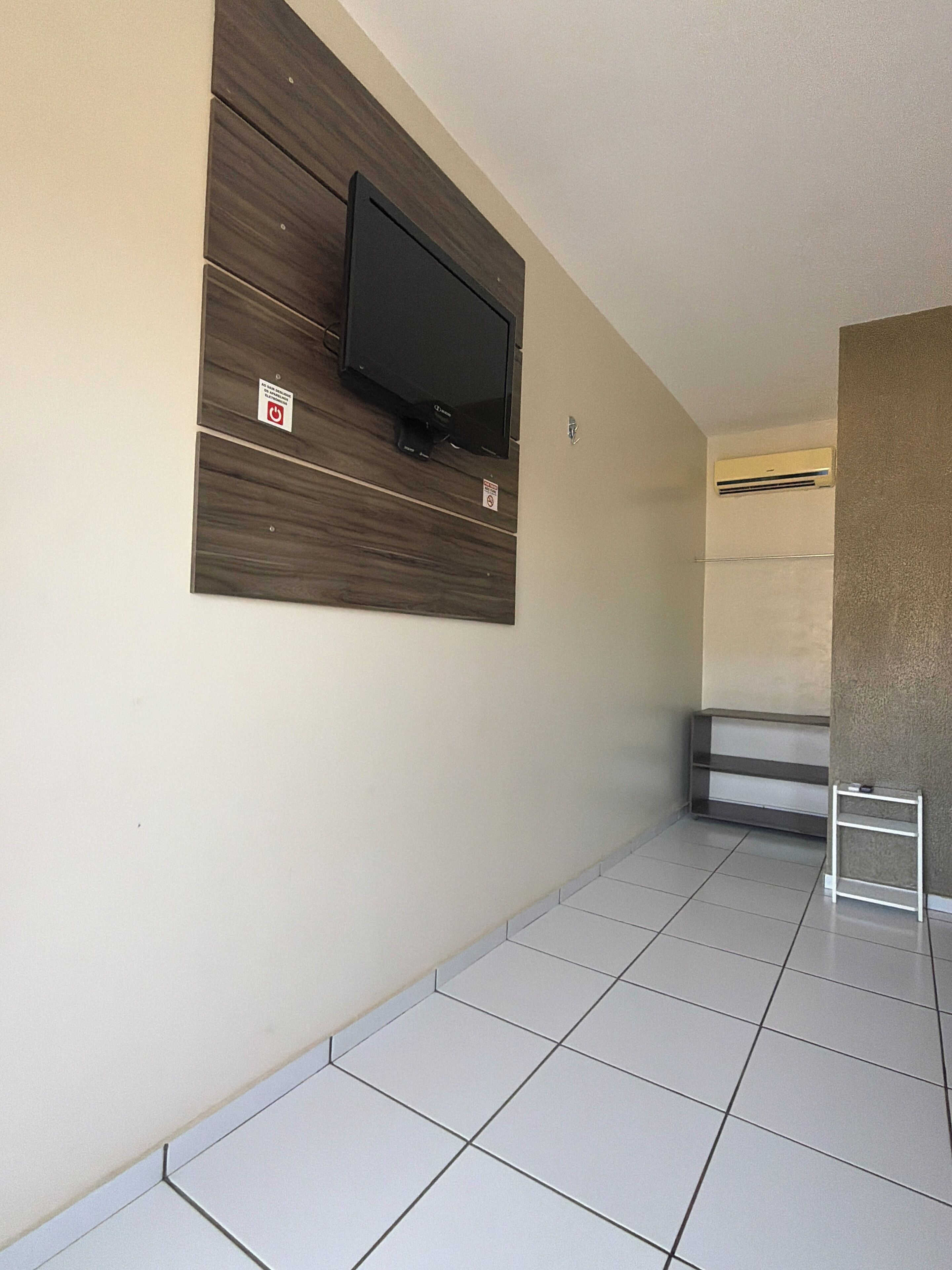 Quarto com televisão de tela plana fixada em parede de painel de madeira, piso ladrilhado e pequena prateleira branca.