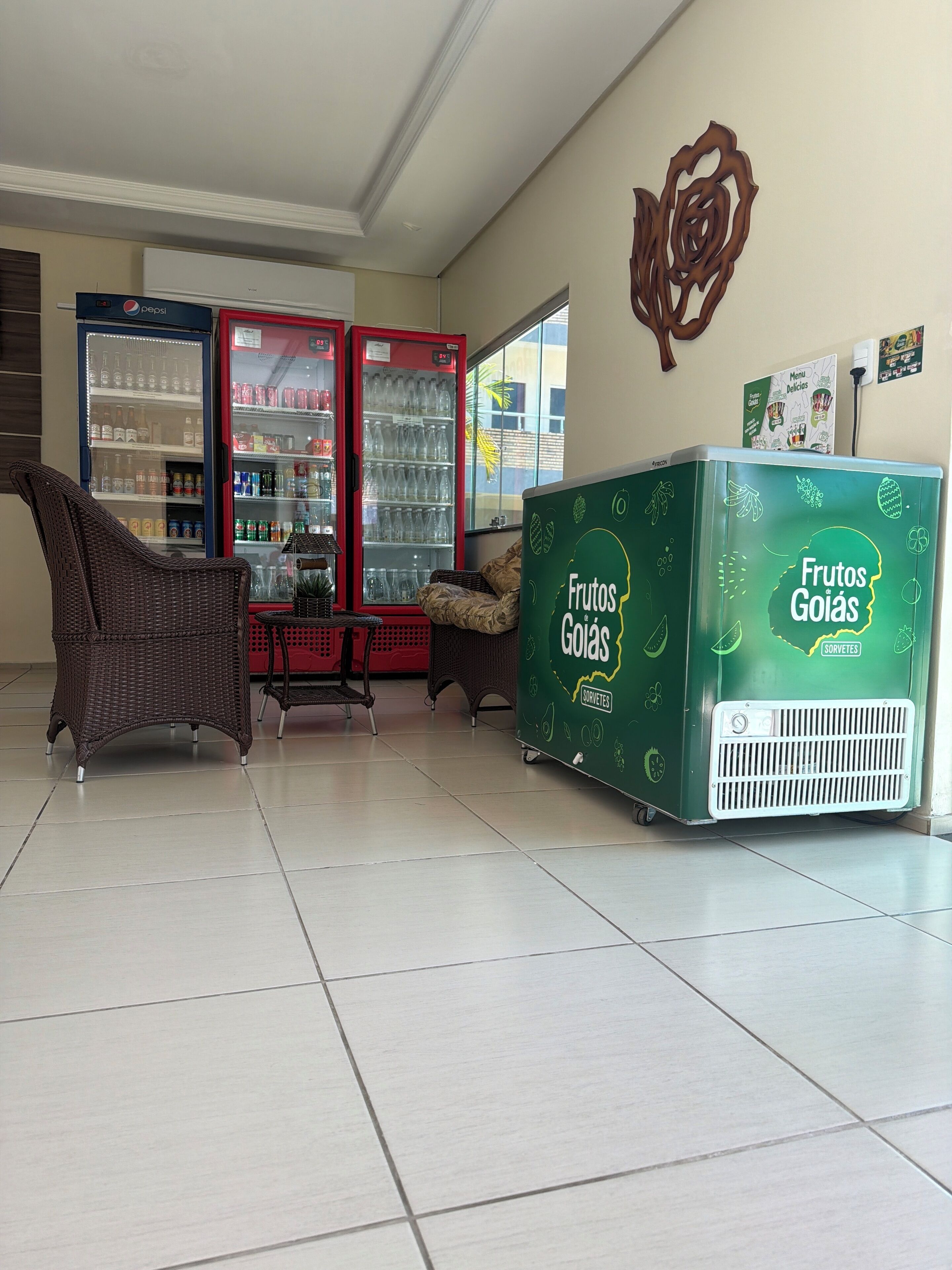 Una habitación con una silla de mimbre, dos refrigeradores llenos de bebidas y un carrito verde con la etiqueta 'Frutos Goías'.