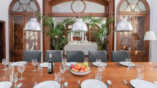 Ein Esszimmer mit einem langen Tisch, der für eine Mahlzeit gedeckt ist, umgeben von Stühlen, mit einer Obstschale und Weingläsern auf dem Tisch.