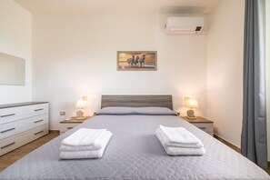 Un lit bien fait, avec des oreillers, deux tables de chevet avec des lampes, un climatiseur fixé au mur et un tableau au-dessus du lit.