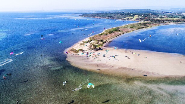 Eine Luftaufnahme eines Strandes mit klarem Wasser, sandigem Ufer und mehreren Kitesurfern.