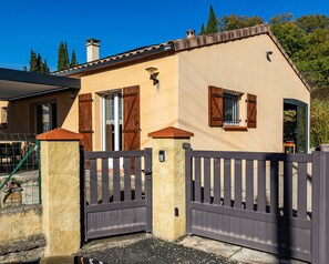 Une maison à l’extérieur beige, avec des volets en bois, une entrée fermée par un portail et un pilier en pierre.