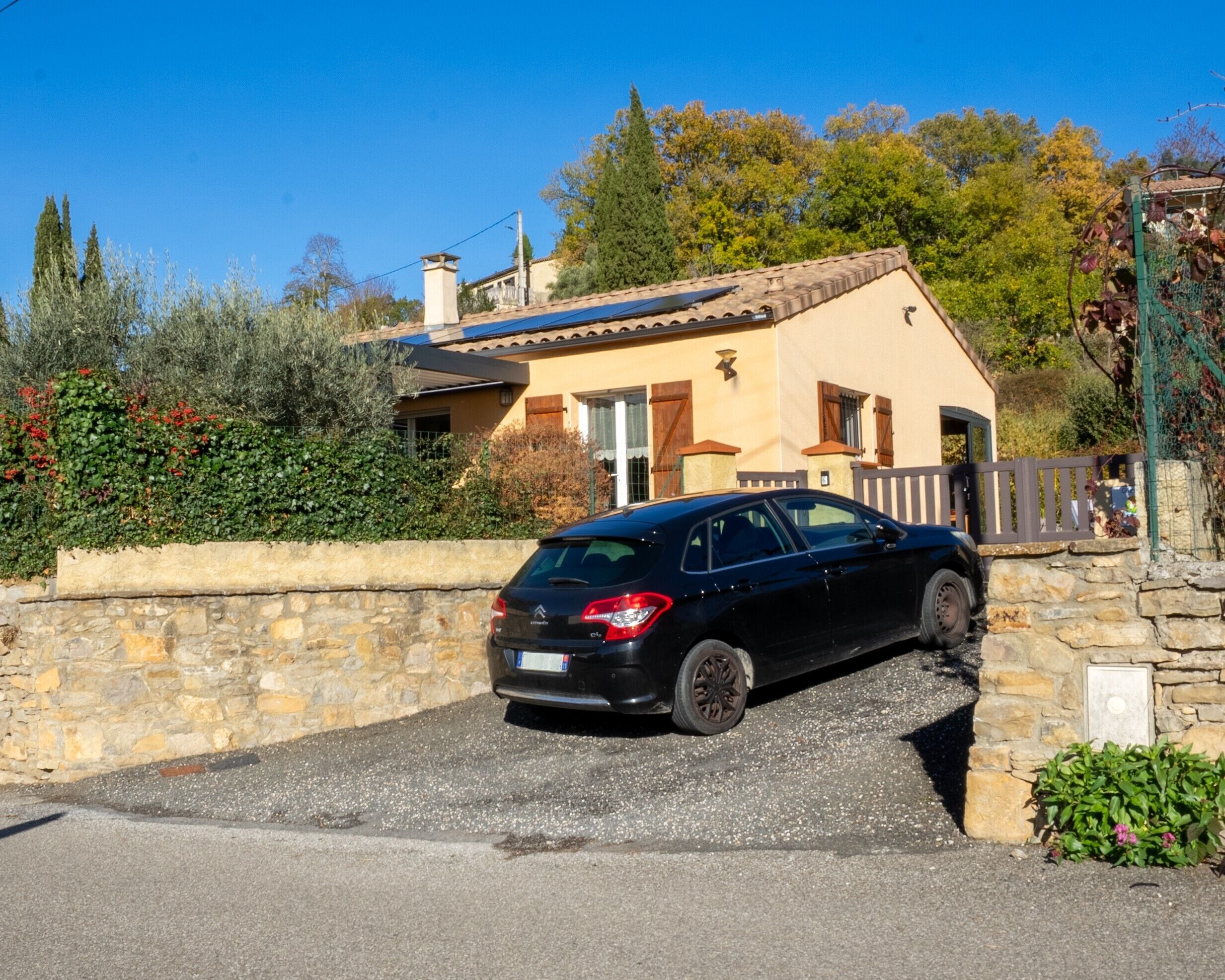 A house with a stone wall, a parked black car, and a clear blue sky.