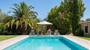 Una piscina con sillas de descanso y una sombrilla, rodeada de palmeras y un edificio de piedra.