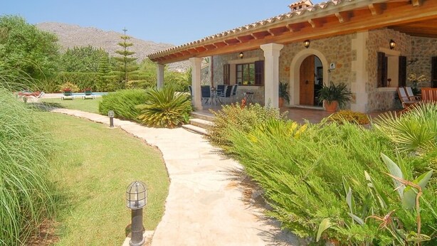 Una casa de fachada de piedra, un porche cubierto con mobiliario para exterior, y un jardín cuidado con diversas plantas y un sendero que lleva a la entrada.