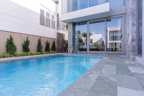 Ett modernt poolområde med en klarblå pool, stenplattor och en glasdörr som leder in till en byggnad med en vit fasad och en balkong.