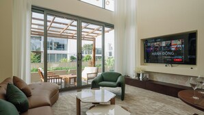 Een moderne woonkamer met een bank, fauteuil en salontafel, een aan de muur bevestigde televisie en een balkon met tuinmeubilair.