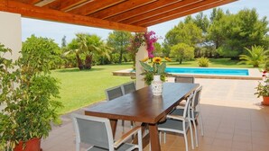 Un pati exterior amb taula i cadires de fusta, un vas amb flors i vista a una piscina i vegetació.