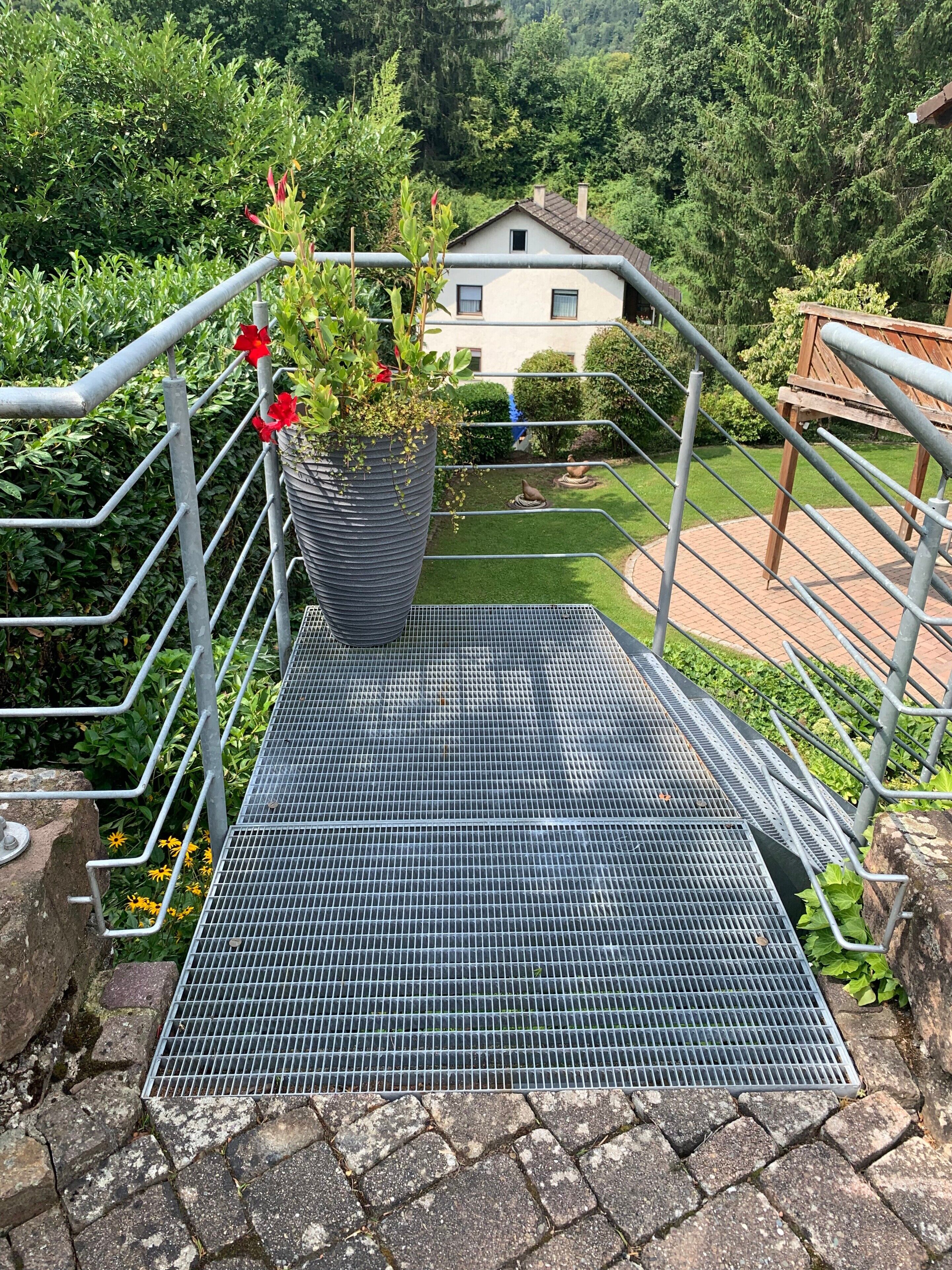 A metal staircase with a metal grid floor leading to a house surrounded by greenery.