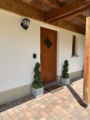 Una porta in legno con una finestra a forma di diamante, due piante in vaso, una piccola finestra e un muro con superficie strutturata.