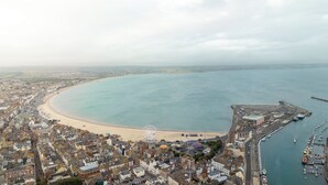Vue aérienne d’une ville côtière avec une plage de sable, une étendue d’eau importante et un port de plaisance rempli de bateaux.