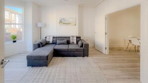 A modern living room with a sectional sofa, a dining area with a table and chairs, a large window with a view of buildings, and a ceiling fan. - Nelson Road - 3 Bed Terrace in Yarmouth (Great Yarmouth)