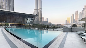 Un espace piscine moderne avec une bordure en verre, entouré d’une terrasse carrelée et de chaises longues, offrant une vue sur un paysage urbain avec de hauts immeubles.