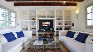 Een woonkamer met witte banken, een glazen salontafel en een televisie op een dressoir. Er staan ook boeken en decoratieve objecten op planken.