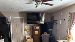 A spacious living room with a ceiling fan, a flat-screen TV, wooden flooring, and a dining area with a table and chairs.