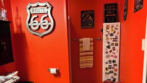 A bathroom with a red wall, a Route 66 sign, a toilet, a towel rack, and a refrigerator covered in magnets.