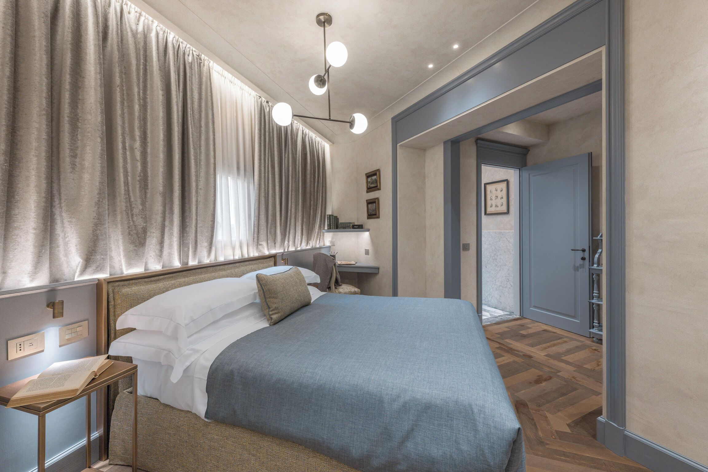 A bedroom with a large bed, a bedside table with a book, a nightstand, and a lamp. There is a door leading to another room.