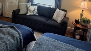 Et værelse med en sort sofa, to senge med blå sengetøj, en loftsvifte, et vindue med blå gardiner, en dør og et maleri på væggen.