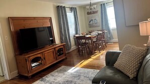 En stue med stort fladskærms-tv, tv-møbel i træ, mørk lædersofa og spiseområde med bord og stole.