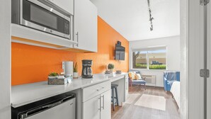 Een moderne keuken met witte kasten, een oranje accentmuur, een magnetron, een koffiezetapparaat en een raam met uitzicht op een parkeergarage.