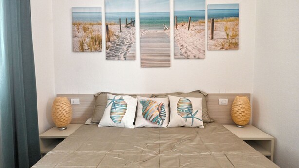 Une chambre à coucher comprenant un lit, une tête de lit, des tables de chevet, des lampes et une œuvre d’art fixée au mur représentant une scène de plage.