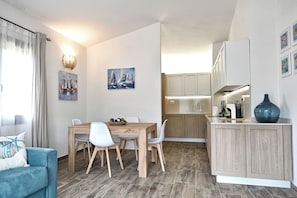 A modern kitchen with wooden cabinets, a dining area with a wooden table and white chairs, and a living area with a blue sofa and a lamp. - Villetta Dalia (San Teodoro)