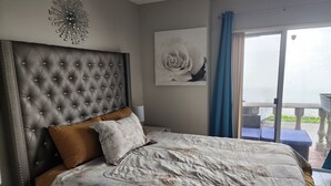Een slaapkamer met een gestoffeerd hoofdeinde, een bed met een beddegoed met patroon, een nachtkastje, een wanddecoratie en uitzicht op het buitenlands landschap door een schuifdeur.
