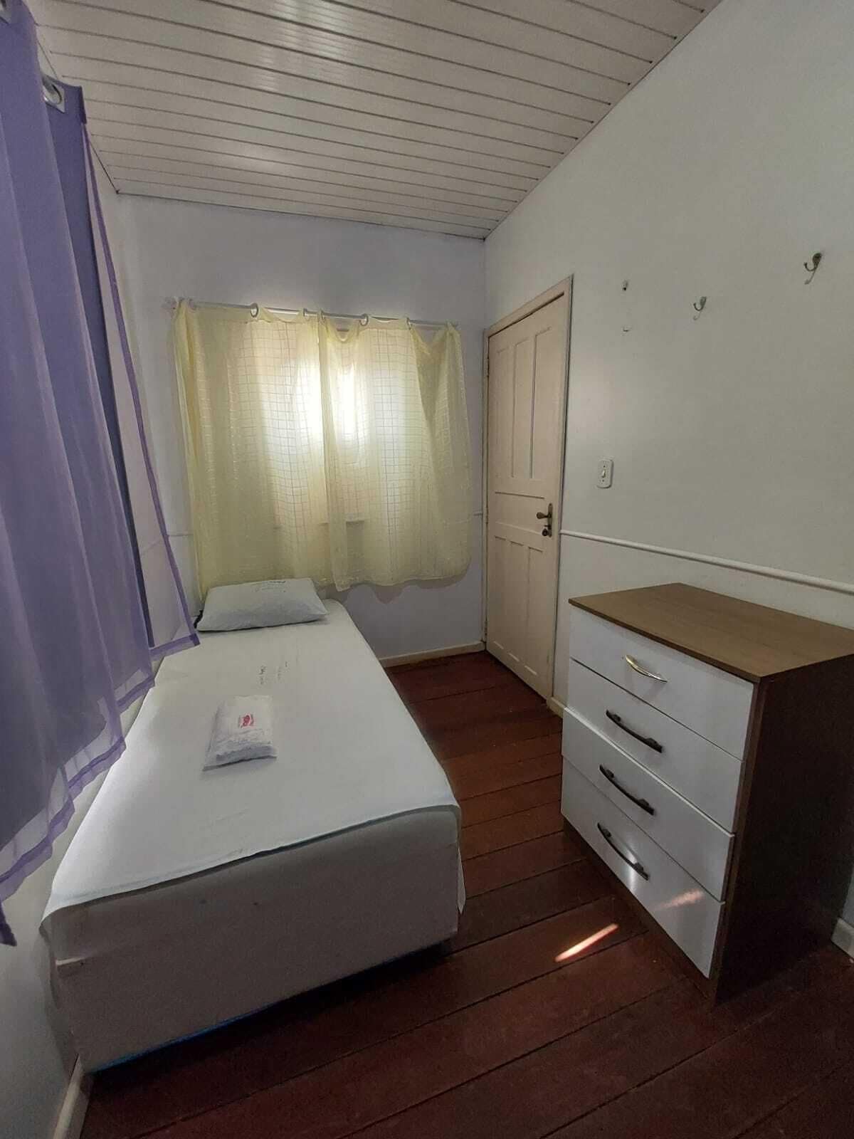A small, single-bed room with a wooden floor, a white bed, a wooden dresser with drawers, a door, and a window with sheer curtains.