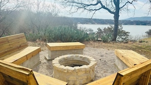 An outdoor seating area with wooden benches and a stone fire pit, surrounded by gravel and cacti, overlooking a lake and distant hills. - Gated Lake Ranch, Private Dock & 3.5 Acres, Fire pit & Pet Friendly (Palo Pinto)